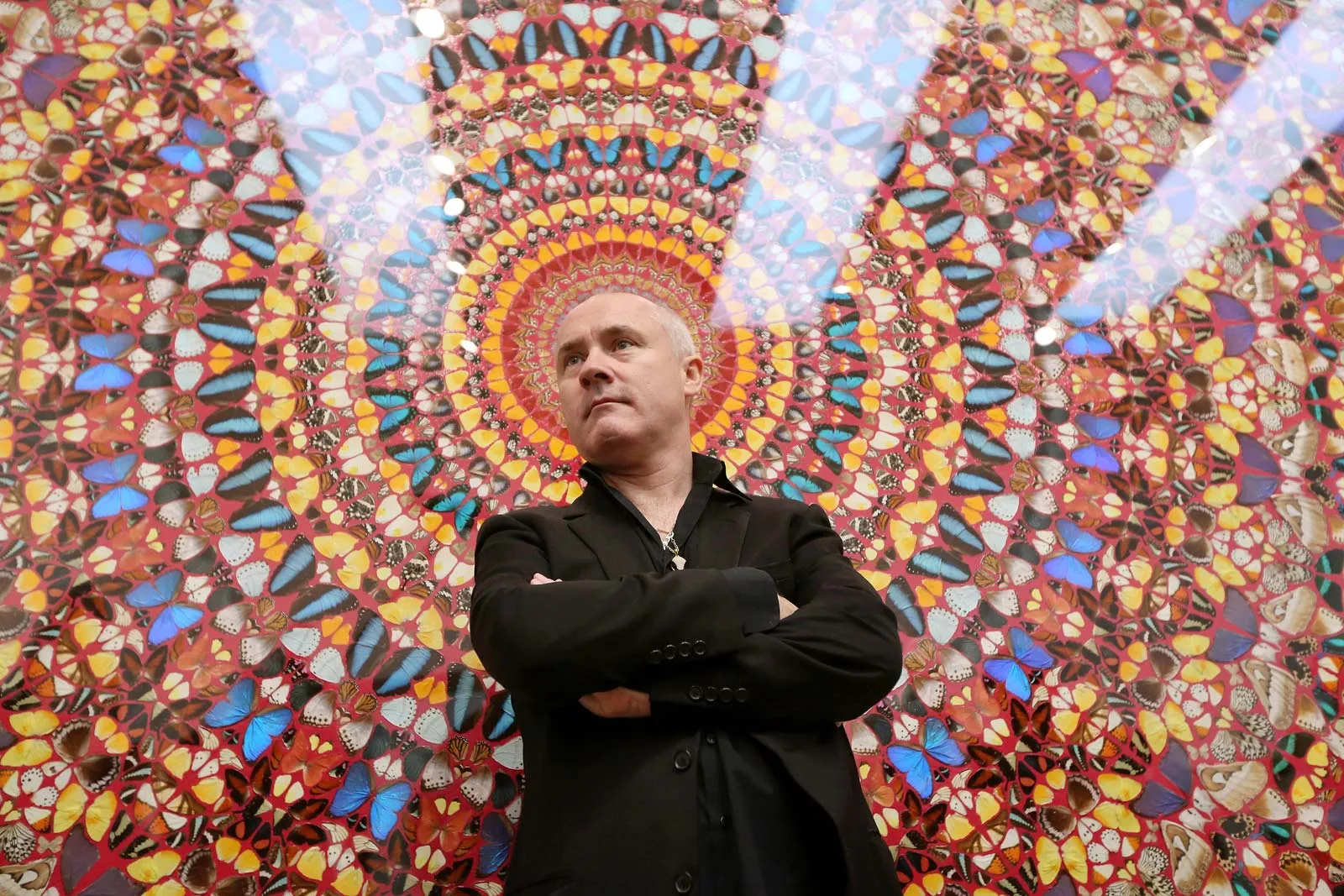 Damien Hirst standing with his arms folded in front of one of his butterfly kaleidoscope paintings, a dense circular mandala of coloured butterfly wings on a deep red ground