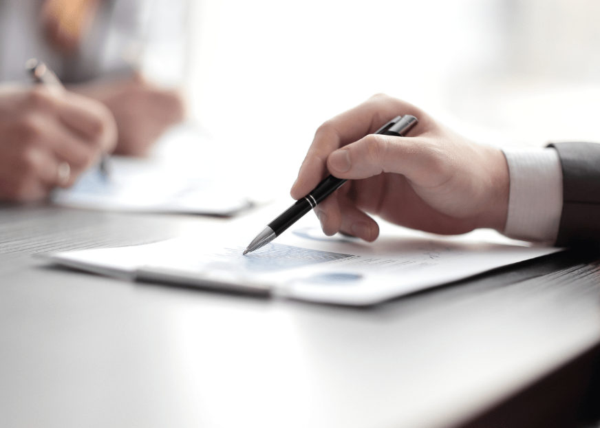 A hand resting the tip of a pen on a printed document of figures and charts on a desk, with a second person writing on their own copy behind