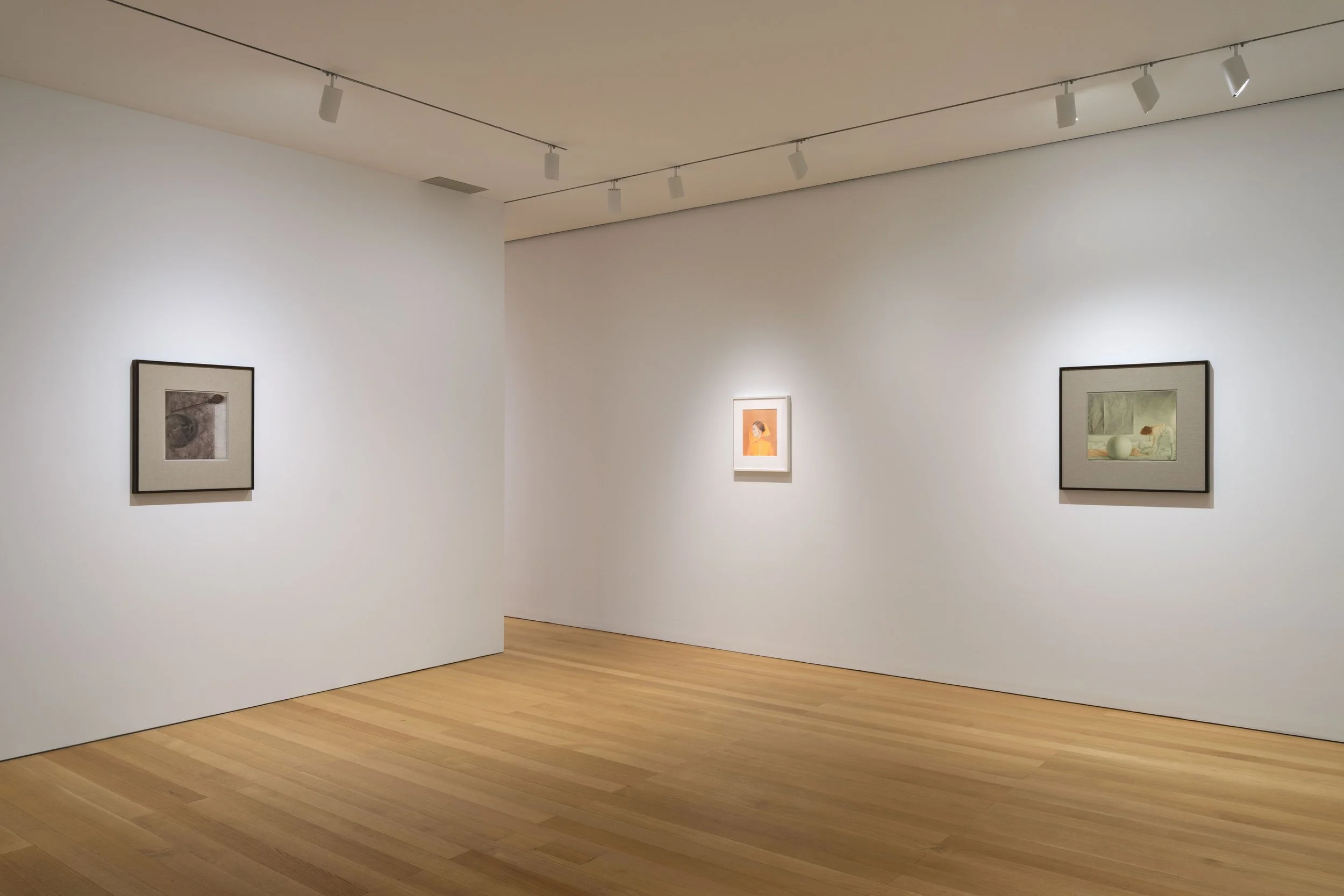 Installation view of a gallery hang: three framed works on white walls under track lighting
