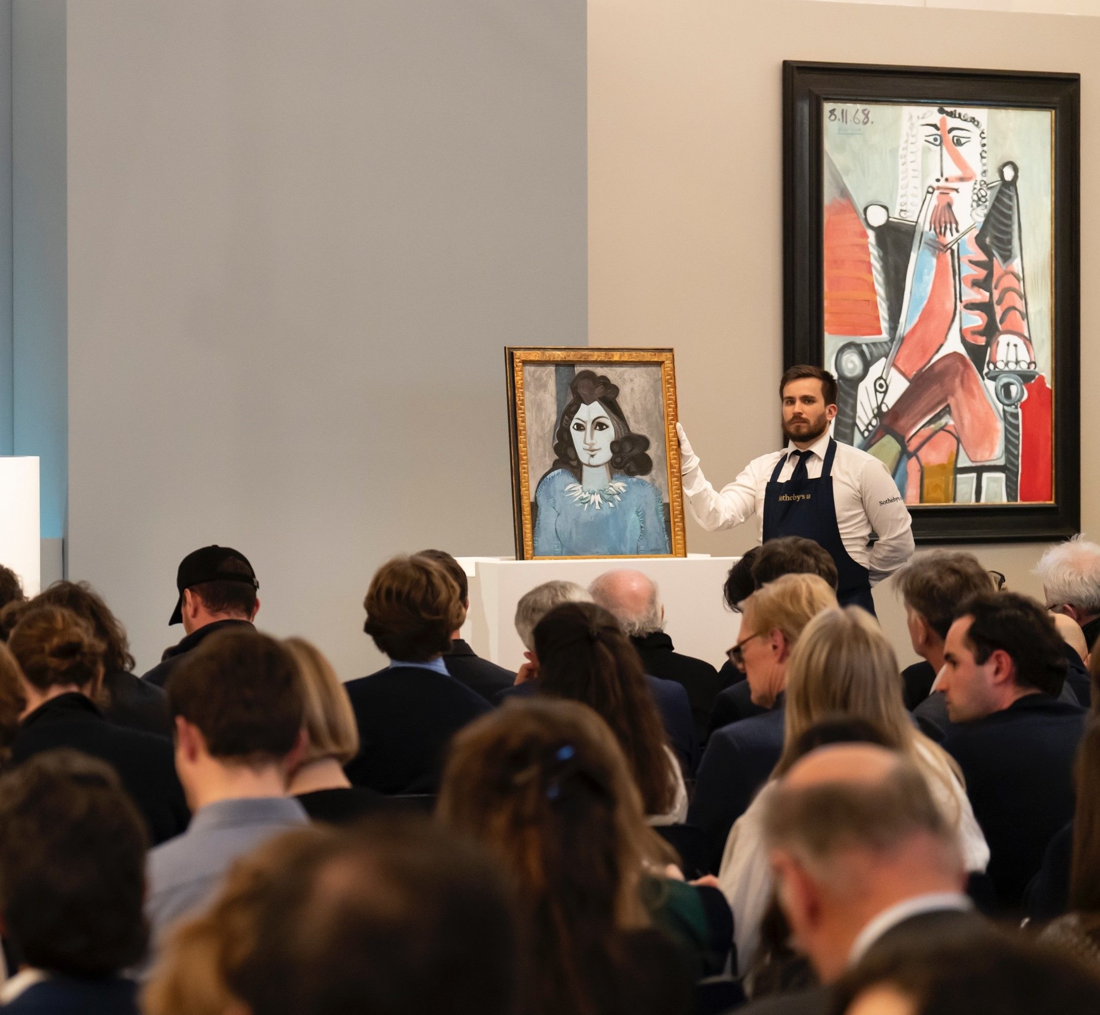 A porter in an apron and white gloves steadying a framed portrait on a plinth in a packed saleroom, with rows of seated bidders in front of him and a large framed painting on the wall behind