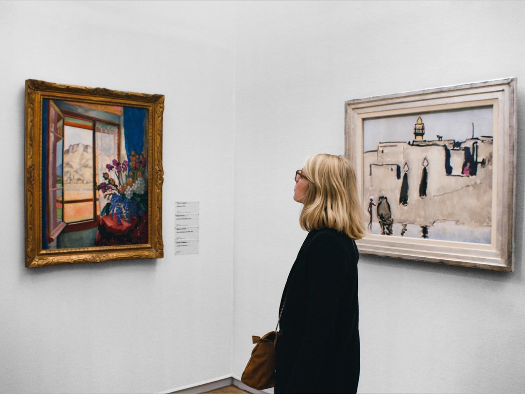 A visitor standing close to a framed painting in a gallery, with the work's printed wall label on the wall beside it
