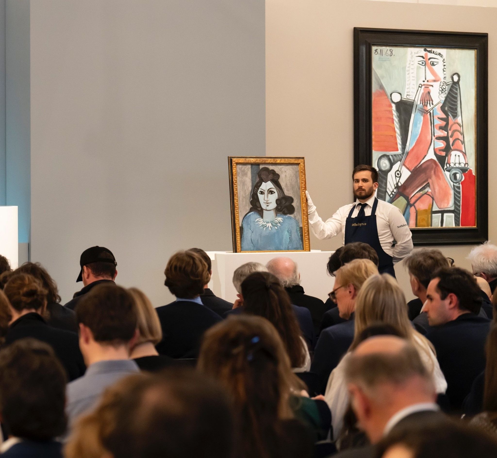 A porter in an apron and white gloves steadying a framed portrait on a plinth in a packed saleroom, with rows of seated bidders in front of him and a large framed painting on the wall behind